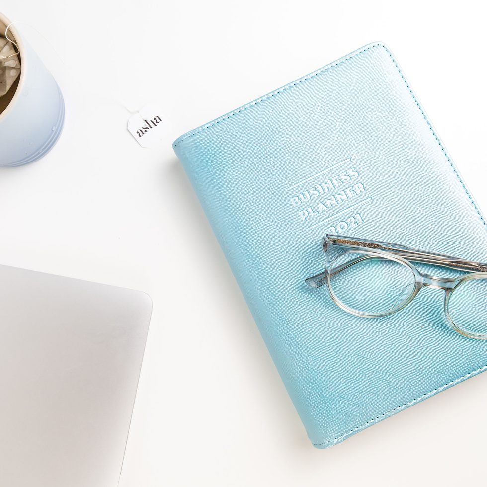 A pale blue 2021 business planner with reading glasses resting on it, beside a laptop and a cup of tea